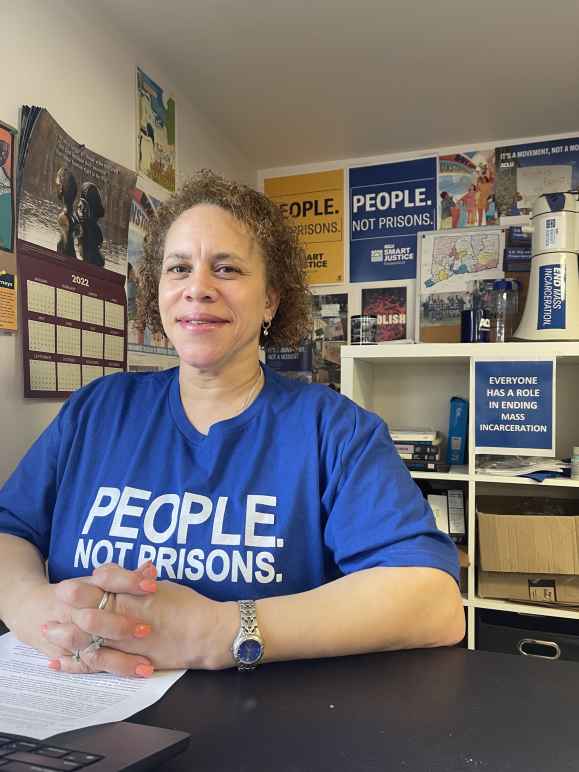 ACLU of Connecticut Smart Justice leader Pierlette Jones sits behind a desk, hands folded, smiling at the camera. She is wearing a blue people not prisons shirt and a watch. A copy of legislative testimony is under her clasped hands. 