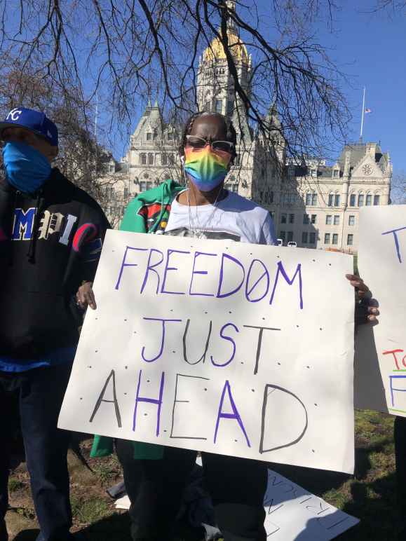 ACLU-CT Smart Justice leader Terri Ricks stands in front of the CT State Capitol Building. She is wearing sunglasses and a rainbow face mask, and she holds a white sign that says, in blue letters, "FREEDOM JUST AHEAD."