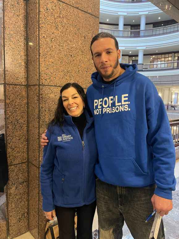 ACLUCT Smart Justice leaders Will and L stand at the capitol, smiling and wearing people not prisons blue hoodies