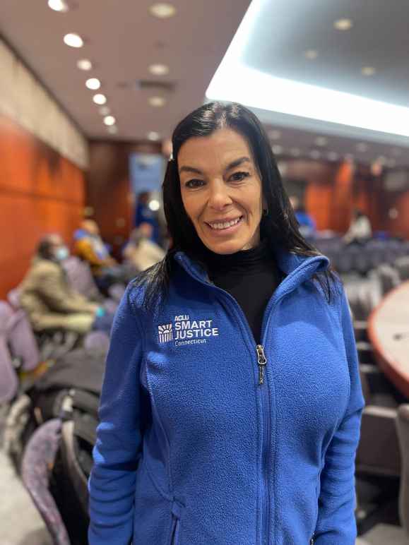 ACLU-CT smart justice leader Lori LeDonne stands, smiling at the camera and wearing a blue zip up sweatshirt. Behind her is a legislative hearing room