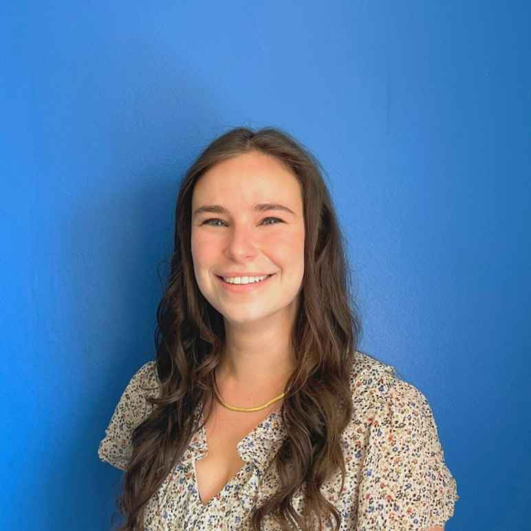 Jess Zaccagnino, a white woman wearing a floral dress with long brown hair past her shoulders, smiling at the camera in front of a blue wall.