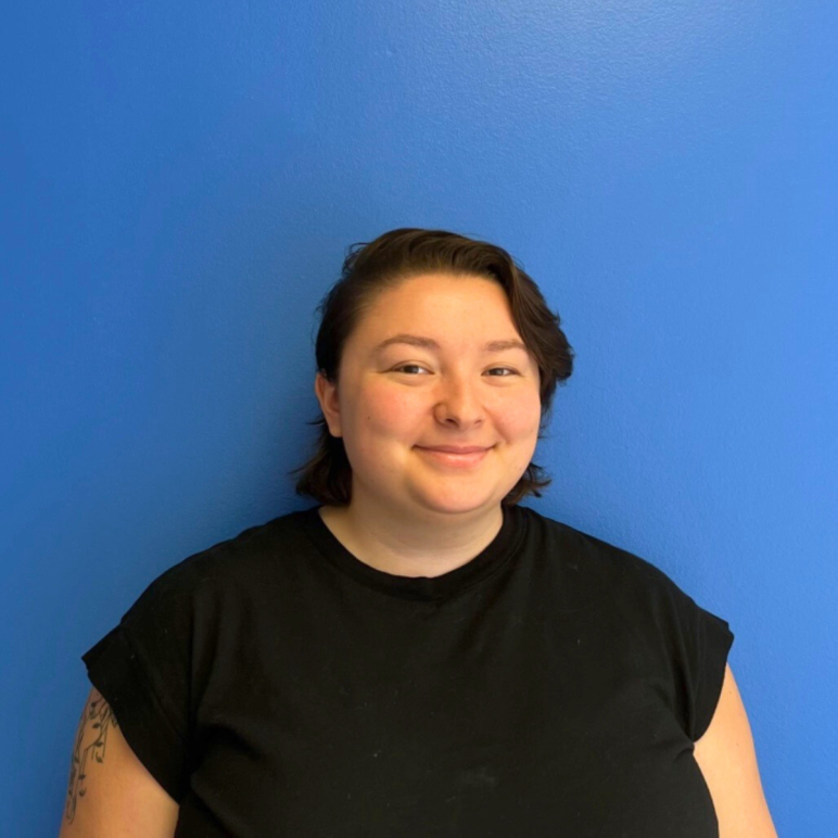 On a blue background, a person looks at the camera with a smile. Kira is wearing a black t-shirt and has short, dark hair with bangs on their left side.