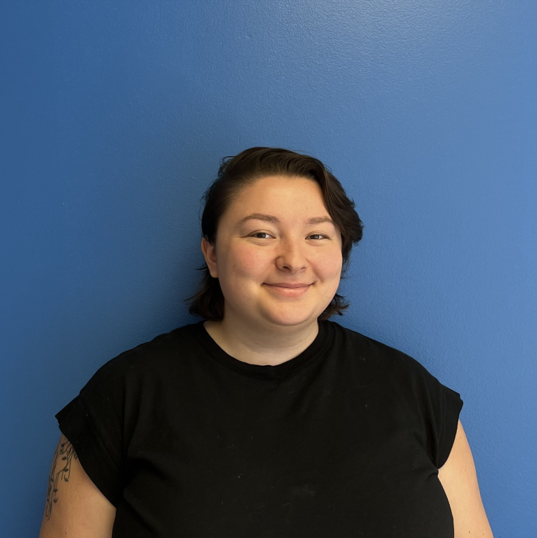 On a blue background, a person looks at the camera with a smile. Kira is wearing a black t-shirt and has short, dark hair with bangs on their left side.