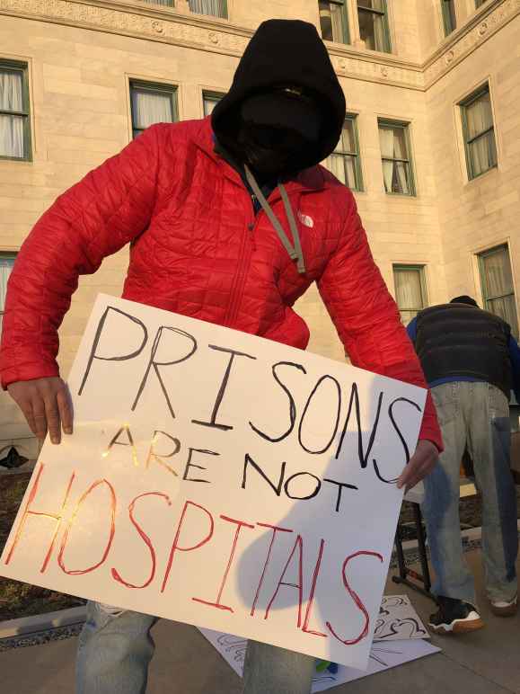 A person in a red coat, black facemask, and black hood holds a sign that says "Prisons are not hospitals" in front of the Connecticut State Capitol building in December 2020.