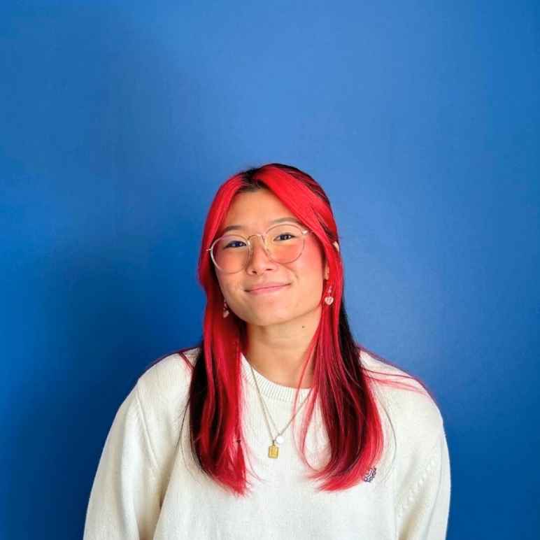 A Korean woman with long black hair, with red dyed hair in the front of and under her black hair is smiling at the camera. She is wearing gold-rim glasses and a white sweater on a blue background.