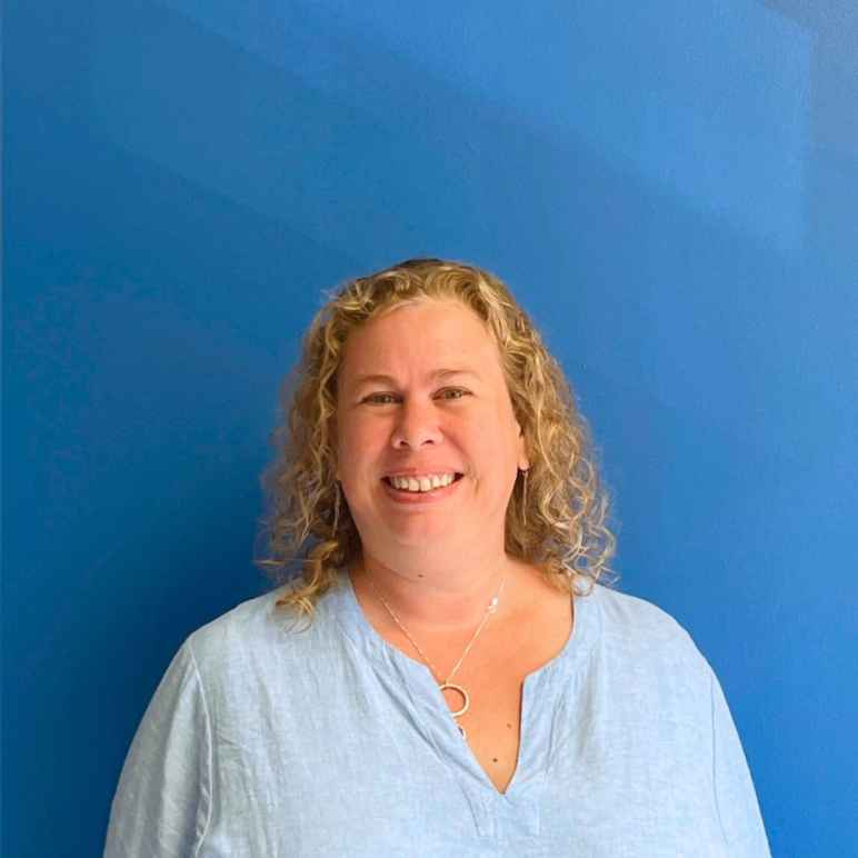 A white woman with curly blonde hair to her shoulders in a collared shirt smiles at the camera in front of a blue wall.