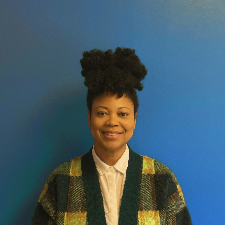 A Black woman with her curls up on top of her head in a green plaid cardigan smiles at the camera in front of a blue wall.