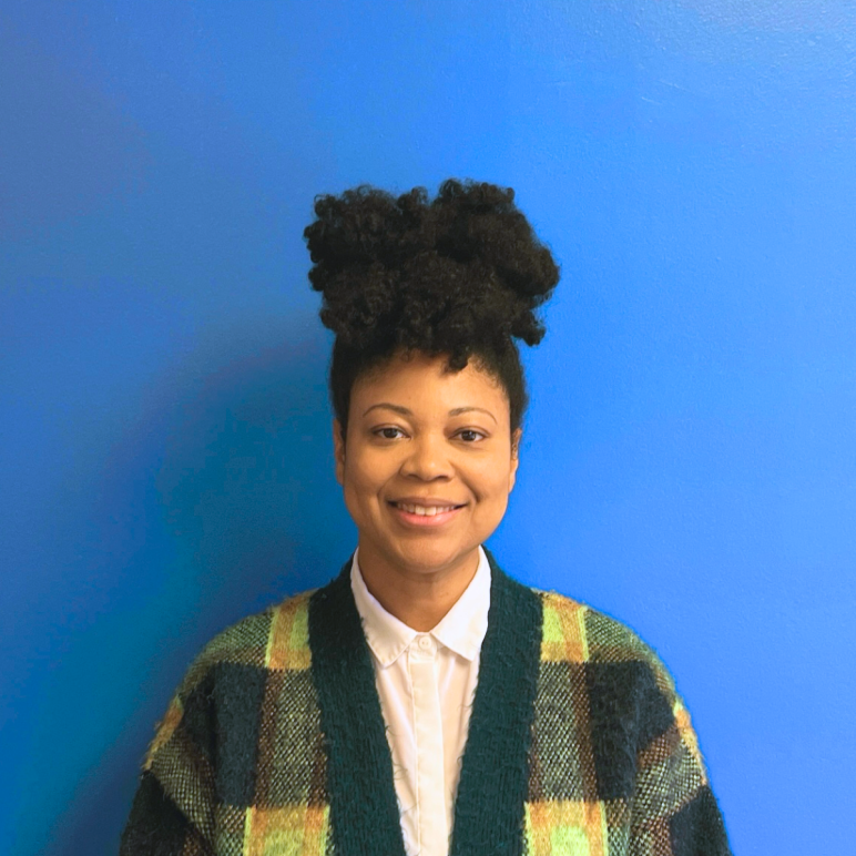 A Black woman with her curls up on top of her head in a green plaid cardigan smiles at the camera in front of a blue wall.