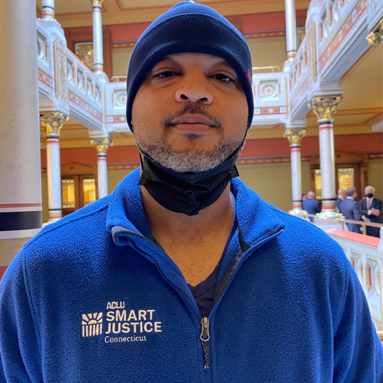 Image description: Tyran Sampson, ACLU-CT Smart Justice leader, stands, facing the camera. He is wearing a blue ACLU of Connecticut Smart Justice zip-up sweatshirt, a navy blue hat, and has a mask pulled down. He looks serious.