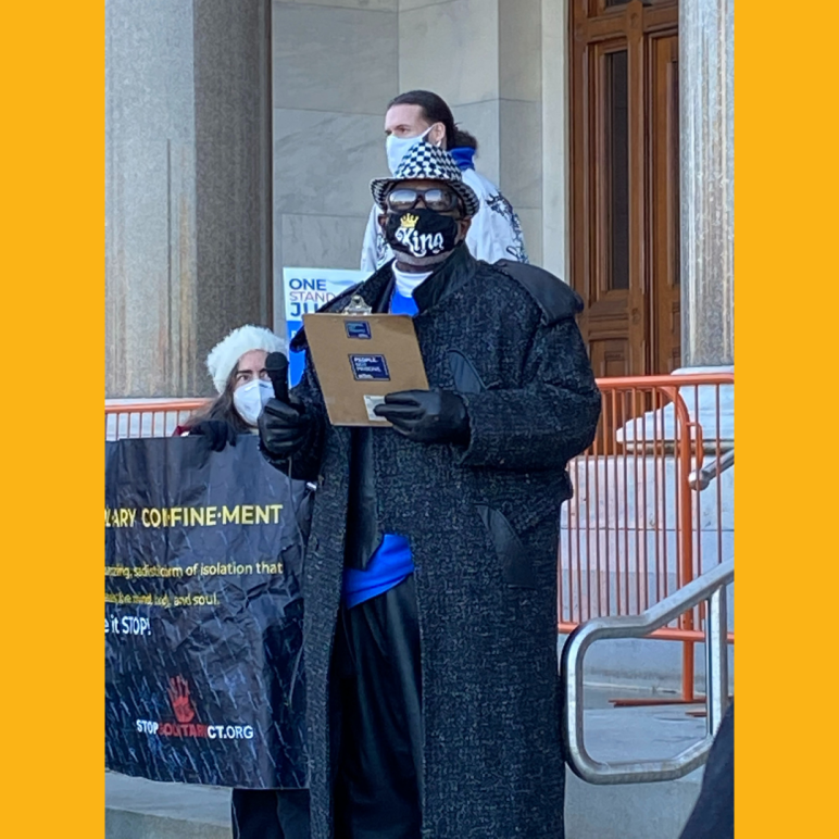 ACLUCT smart justice leader Curtis B. Hudson stands in front of the CT Capitol steps. He is wearing a black and white fedora, sunglasses, black mask, black trenchcoat, and blue Smart Justice shirt, and black gloves, and he is holding a clipboard. 