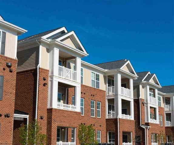 A block of apartments, white on top with balconies and brick below, stand in front of a blue sky.