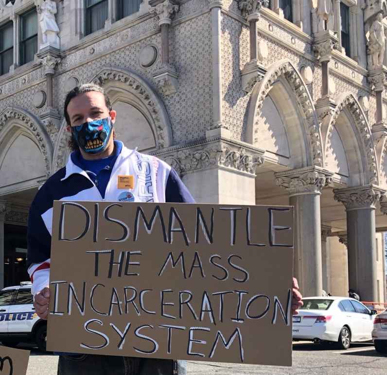 ACLUCT smart justice leader Will stands with a cardboard sign that says, in black and white, "dismantle the mass incarceration system." Behind him is the CT Capitol. he is wearing a mask and a sweatshirt, and yellow people not prison pin