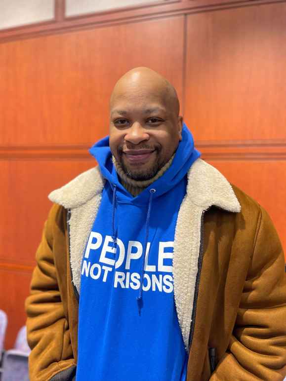 ACLU of CT smart justice leader Donald Rivers smiles, facing the camera and wearing a brown coat over a blue people not prisons hoodie