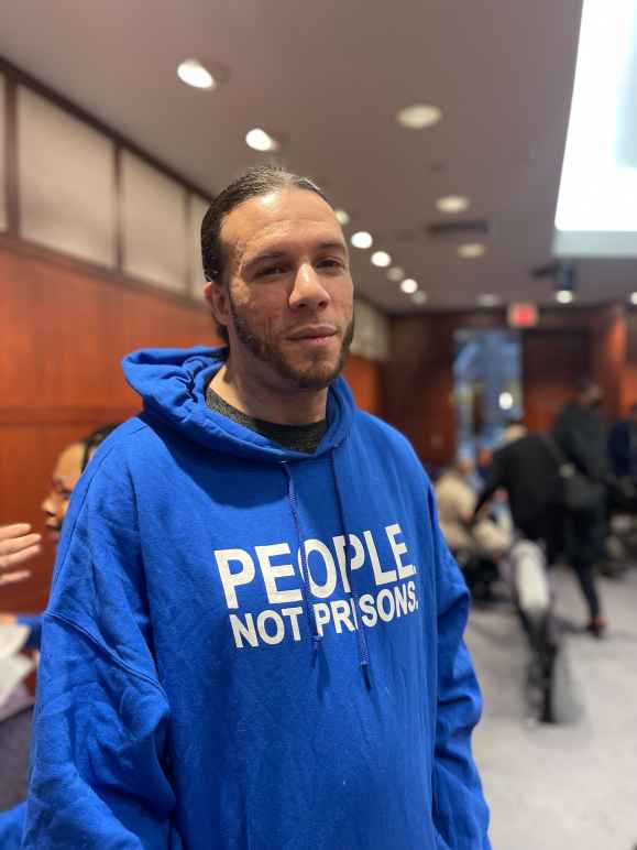 ACLU of CT Smart Justice leader Will Roberts stands, facing the camera, wearing a blue people not prisons hoodie