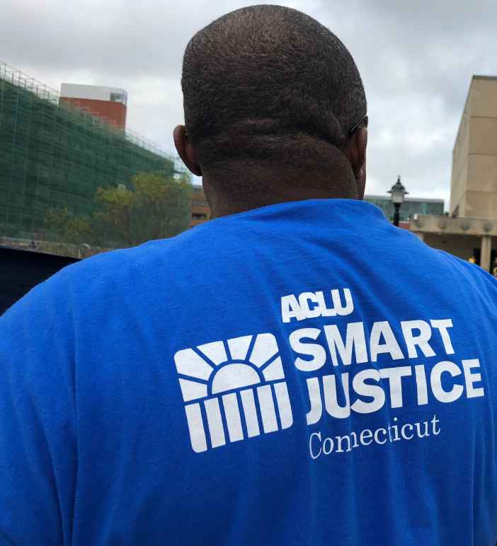 From behind, a man stands in a blue ACLU of Connecticut Smart Justice shirt