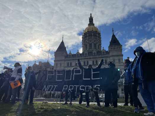 Smart Justice leaders stand in front of the CT capitol. The sun is breaking through a cloud. One person stands with arms upstretched above their head, behind the people not prisons banner