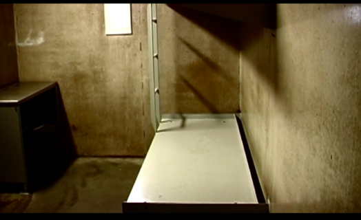 A photo of a bed and desk in a cell at Northern Correctional Institution in Somers, Connecticut. A metal cot-like bed is against a concrete wall on the right. On the left, a small desk and door. The space is small, barren, concrete.