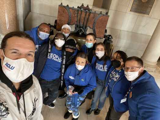 ACLU-CT Smart Justice leaders crouch in front of the bell at the CT State Capitol. They are wearing masks and blue people not prisons shirts, and smiling at the camera.