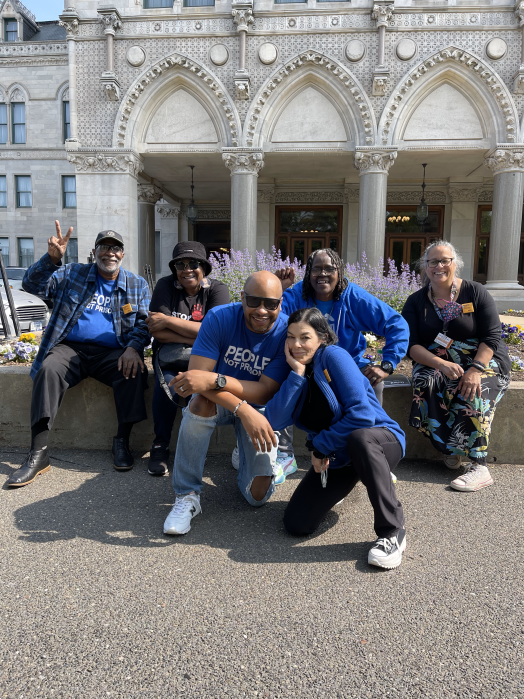 Six ACLU-CT Smart Justice leaders and friends sit and crouch in front of the CT Capitol building. They are smiling, wearing blue "people not prisons" shirts, and the sun is shining.