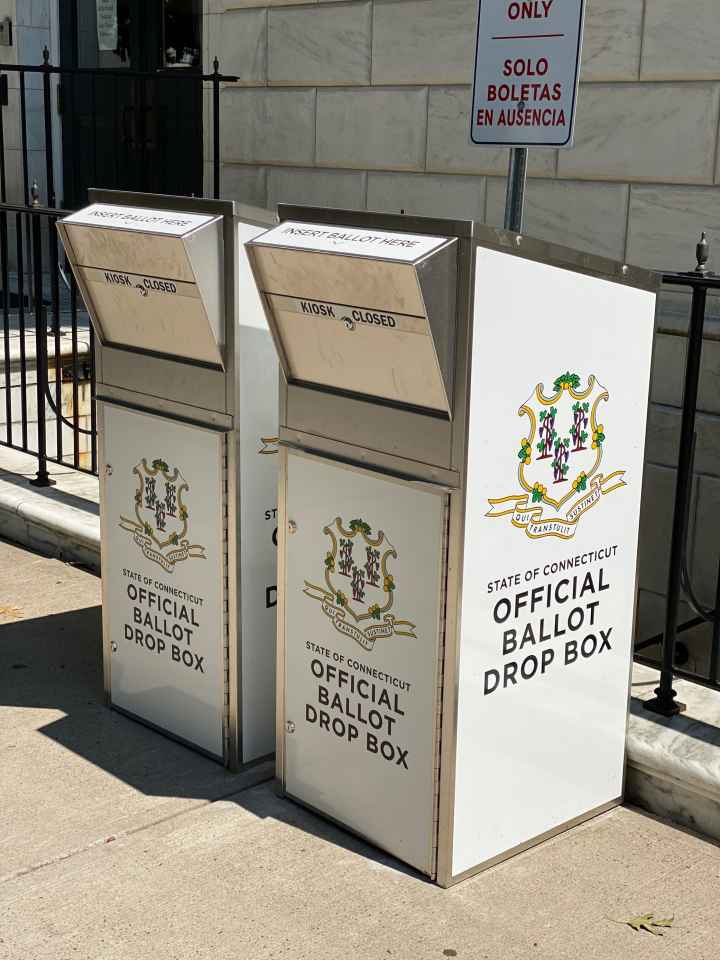 Two official Connecticut ballot drop boxes sit outside of the Waterbury city hall in 2020. The boxes are large, rectangular, and look like library book drops, with the state of CT seal visible, and "official ballot drop box," on their fronts and sides