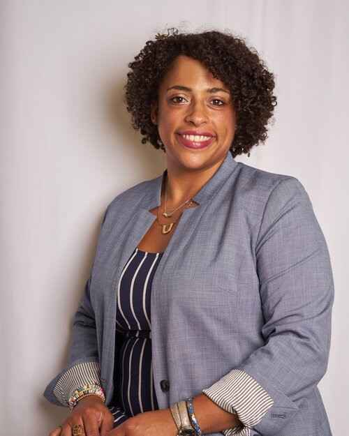 A Black women wearing a grey jacket and vertical striped shirt in front of a white background. The woman is smiling in the image. 