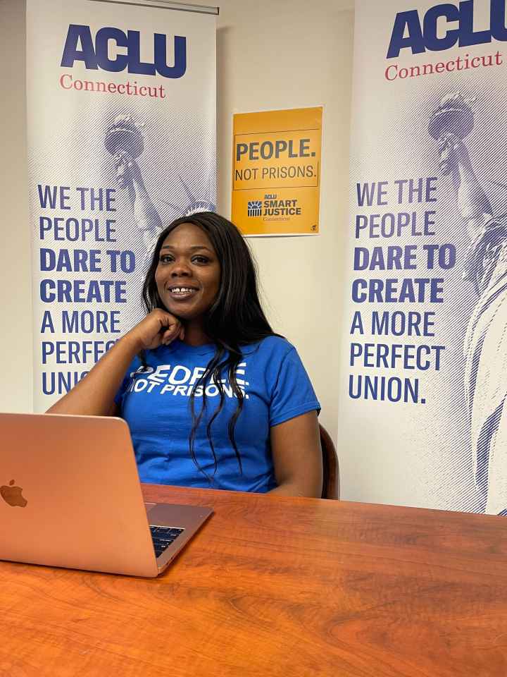 ACLUCT Smart Justice leader Shelby Henderson sits behind a desk, smiling at the camera, with one hand on her chin. She is behind a desk, laptop and wearing a blue people not prisons shirt, with ACLU of CT banners and a people not prisons poster behind her