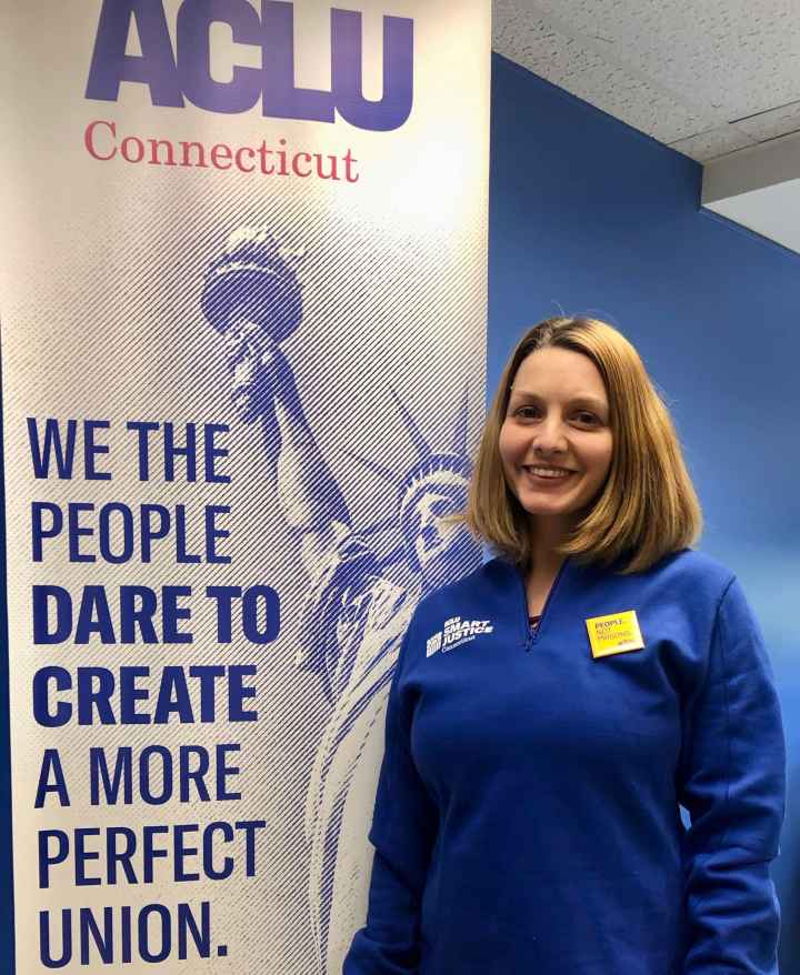 ACLUCT smart justice leader Tracie Bernardi stands facing the camera and smiling. She is wearing a blue Smart Justice zip-up and a yellow People Not Prisons pin. To her left is an ACLUCT banner that says we the people dare to create a more perfect union