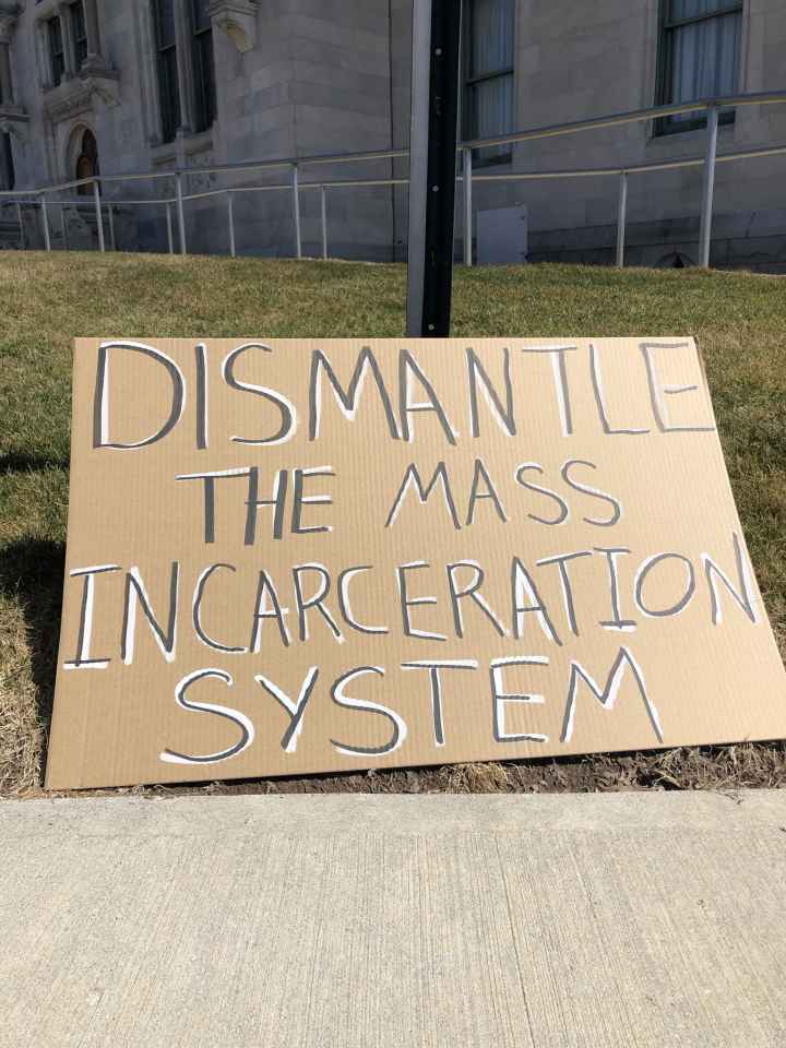 A cardboard sign sits on the grass in front of the CT state capitol building. In black letters with white background, the sign says: "dismantle the mass incarceration system"