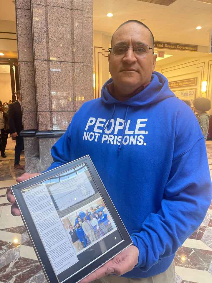 ACLU of CT Smart Justice leader Manuel Sandoval faces the camera, standing and holding a framed collage commemorating his LCSW. he is wearing glasses and a blue people not prisons hoodie