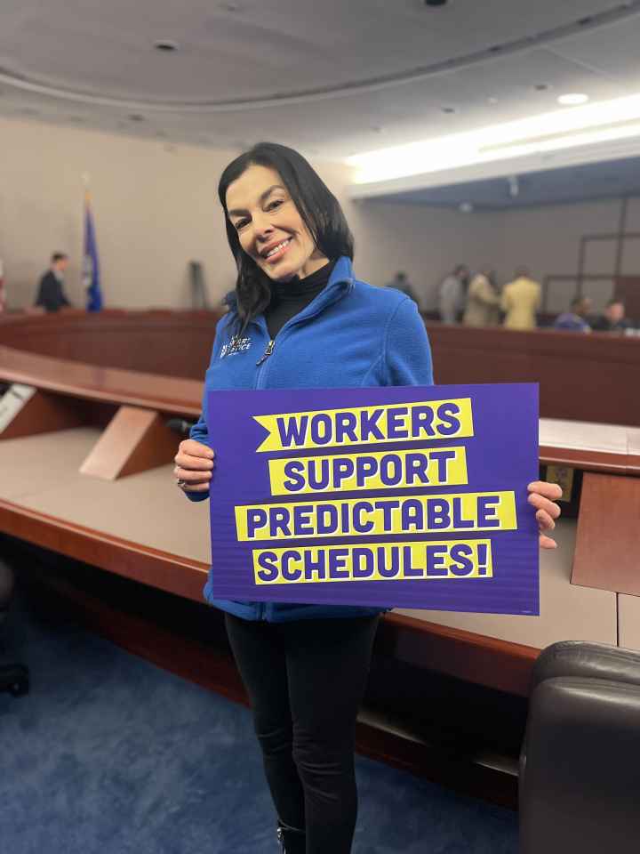 ACLUCT Smart Justice leader Lori LeDonne stands, smiling and holding a "workers support predictable schedules" sign. She is wearing a blue people not prisons zip up sweatshirt