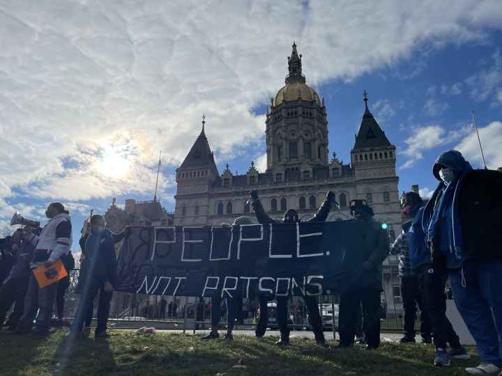 Smart Justice leaders stand in front of the CT capitol. The sun is breaking through a cloud. One person stands with arms upstretched above their head, behind the people not prisons banner