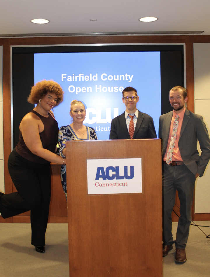 Behind a wooden podium with the ACLU-CT's logo, stands a Black-Taino women with a light brown afro on the left, a white woman with blonde hair to her right, a white man with dark hair and glasses to her right, and a white man with brown hair on the right.
