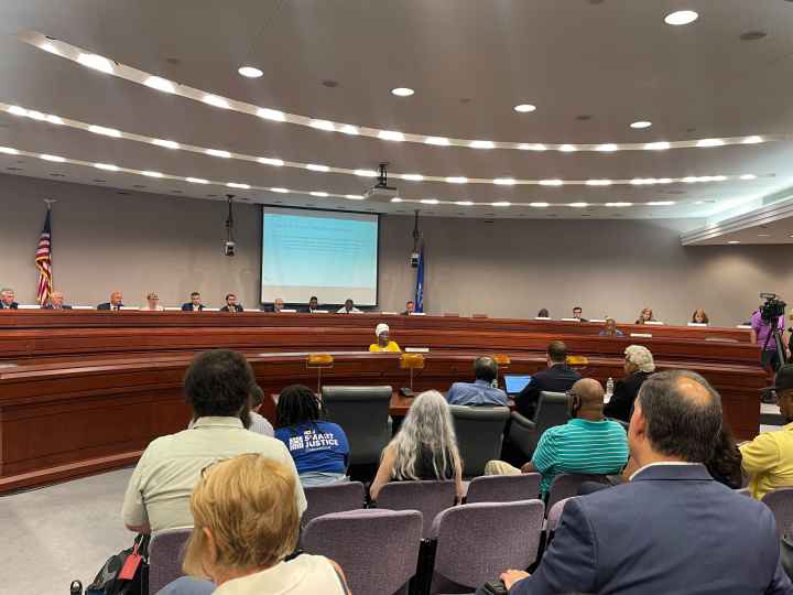 This photo captures a legislative forum about the fake traffic ticket scandal from the back of the room. Several people's backs are towards the camera with legislators in the background of the photo.