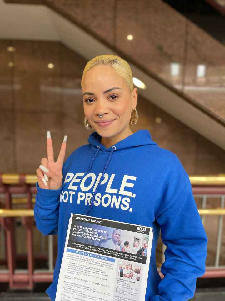 ACLU of CT Smart Justice leader Marquita Reale stands, smiling at the camera and holding up a peace sign. she is wearing a blue people not prisons hoodie