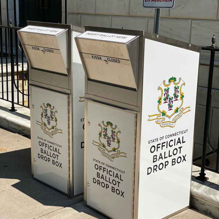 Two official Connecticut ballot drop boxes sit outside of the Waterbury city hall in 2020. The boxes are large, rectangular, and look like library book drops, with the state of CT seal visible, and "official ballot drop box," on their fronts and sides