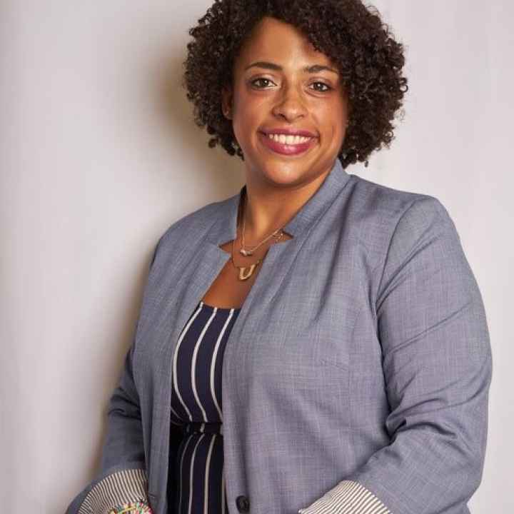 A Black women wearing a grey jacket and vertical striped shirt in front of a white background. The woman is smiling in the image. 