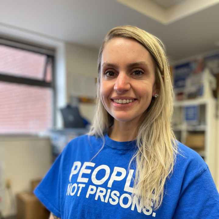 ACLUCT smart justice leader Brittany Lamar stands, smiling and facing the camera. She is wearing a blue people not prisons t-shirt and has long blond hair. 