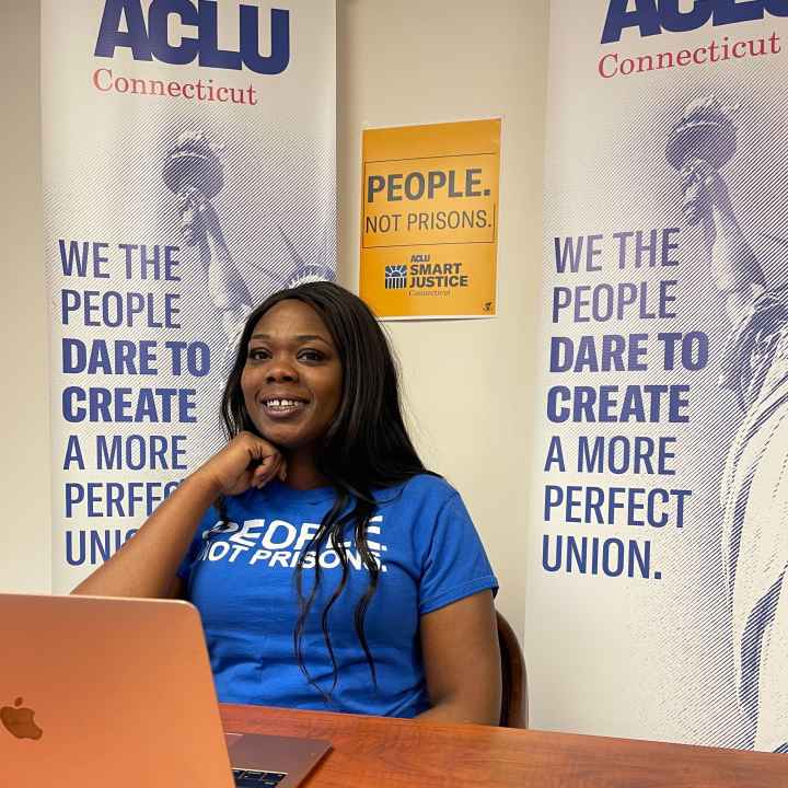ACLUCT Smart Justice leader Shelby Henderson sits behind a desk, smiling at the camera, with one hand on her chin. She is behind a desk, laptop and wearing a blue people not prisons shirt, with ACLU of CT banners and a people not prisons poster behind her