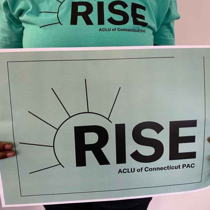 A person with long nails and a ring holds an ACLU-CT Rise PAC sign and wears an ACLU-CT Rise PAC shirt. The shirt and sign are statute of liberty green, the ACLUCT Rise PAC logos in black