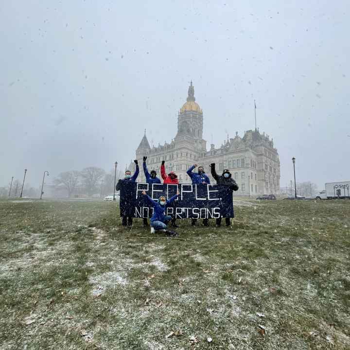 A photo of the Connecticut State Capitol and ACLU-CT Smart Justice leaders with a blue "people not prisons banner." Foreground: capitol lawn, dusted w snow. Middleground: 6 leaders, fists up, hold the banner. Background: snowy sky and capitol building