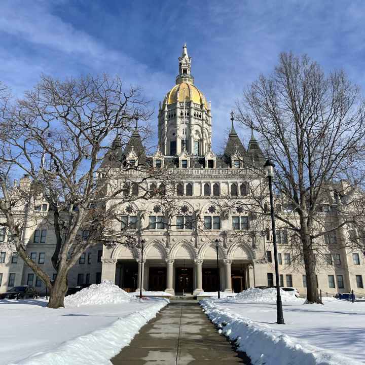 The Connecticut capitol building is centered. The capitol lawn is covered in snow, and a sidewalk leading to the building is clear of snow. The sky is blue and has light clouds.