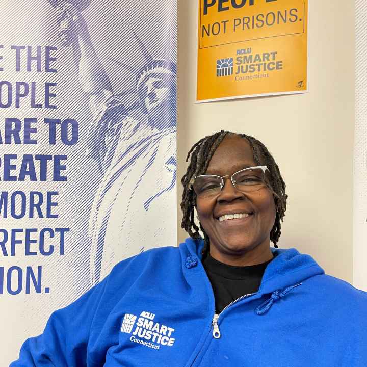 ACLU of CT Smart Justice leader Terri Ricks sits, smiling and facing the camera. She is wearing glasses and a blue people not prisons sweatshirt, in front of a yellow people not prisons sign.