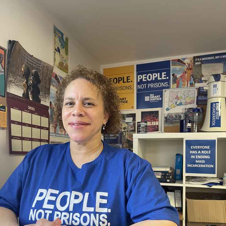 ACLU of Connecticut Smart Justice leader Pierlette Jones sits behind a desk, hands folded, smiling at the camera. She is wearing a blue people not prisons shirt and a watch. A copy of legislative testimony is under her clasped hands. 