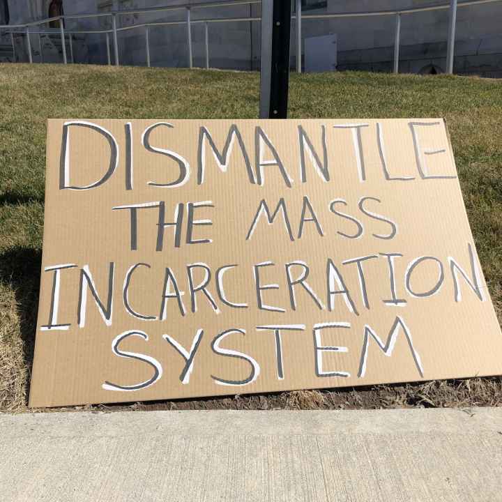 A cardboard sign sits on the grass in front of the CT state capitol building. In black letters with white background, the sign says: "dismantle the mass incarceration system"