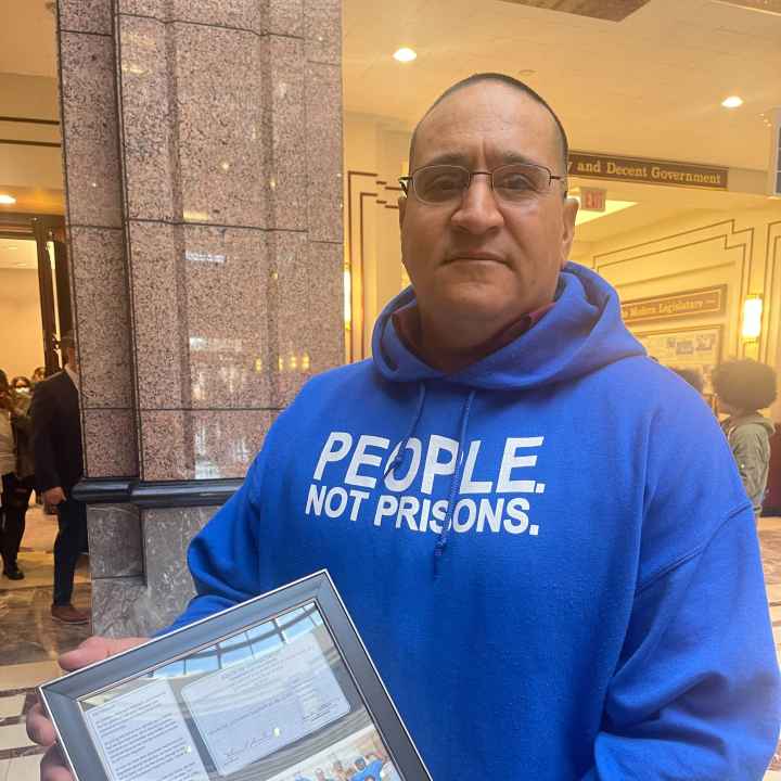 ACLU of CT Smart Justice leader Manuel Sandoval faces the camera, standing and holding a framed collage commemorating his LCSW. he is wearing glasses and a blue people not prisons hoodie