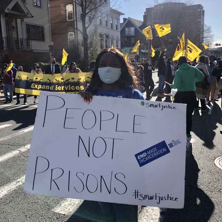 Smart Justice leader Shelby Henderson holds a white sign that says "PEOPLE NOT PRISONS" in blue ink. Behind her is a crowd waving yellow flags and carrying a banner. They are on a street in Hartford, marching.