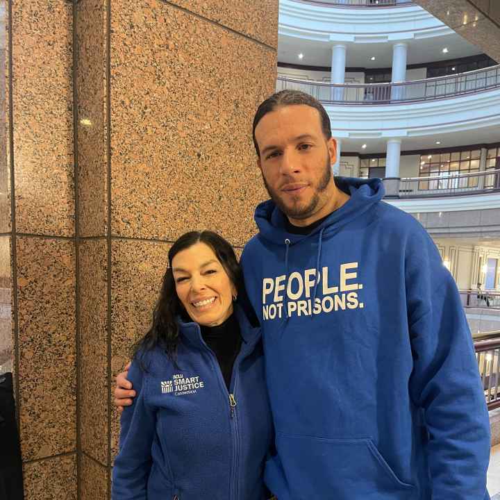 ACLUCT Smart Justice leaders Will and L stand at the capitol, smiling and wearing people not prisons blue hoodies