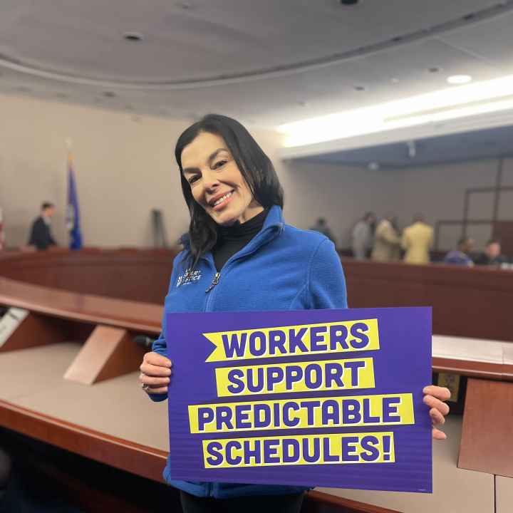 ACLUCT Smart Justice leader Lori LeDonne stands, smiling and holding a "workers support predictable schedules" sign. She is wearing a blue people not prisons zip up sweatshirt