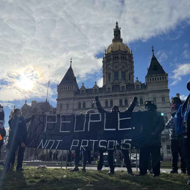 Smart Justice leaders stand in front of the CT capitol. The sun is breaking through a cloud. One person stands with arms upstretched above their head, behind the people not prisons banner