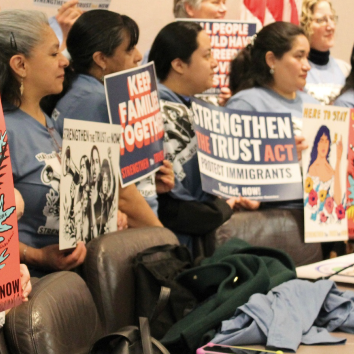 A group of women are standing in a hearing room at the Capitol, holding signs that says "Undocumented + Unafraid" and "Strengthen the Trust Act Now!"